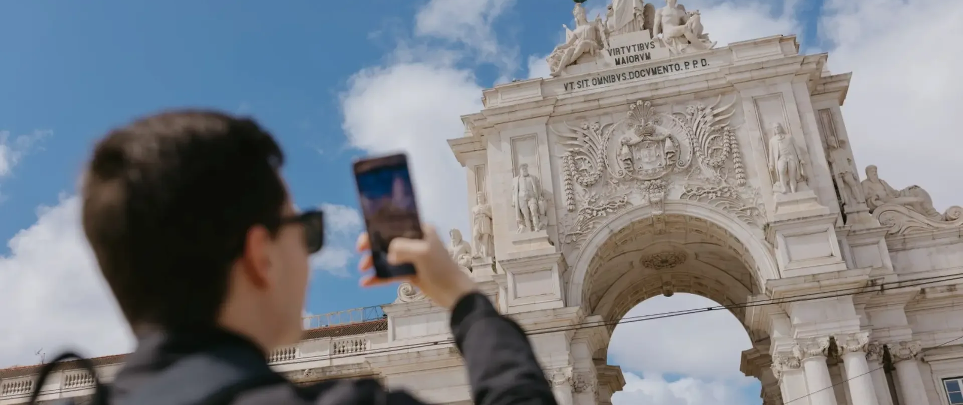 Tourist photographing the Arco da Rua Augusta in Lisbon, Portugal, with clear blue sky and classical sculptures in view.