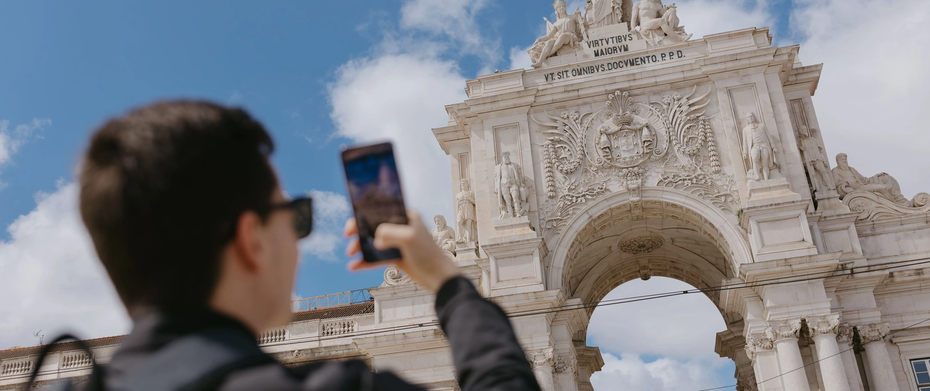 Young tourist takes a smartphone photo of the Rua Augusta Arch in Lisbon, Portugal. The iconic triumphal arch stands against a blue sky with ornate sculptures and historic architecture.