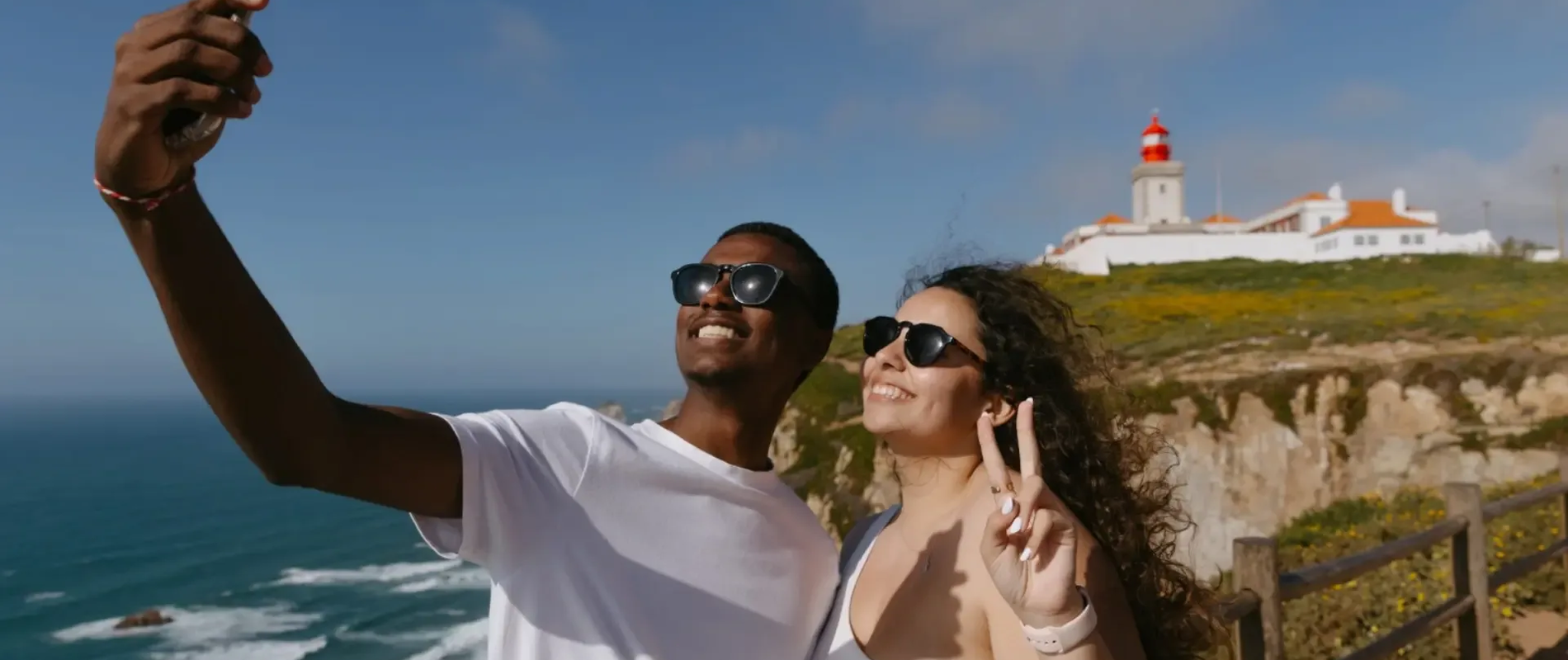 Smiling couple taking a selfie at Cabo da Roca, the westernmost point of mainland Europe, with the iconic red lighthouse in the background.