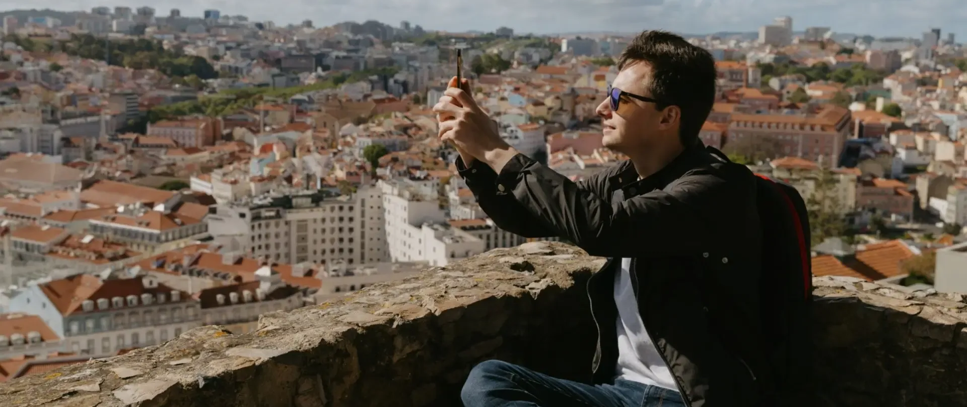 Tourist taking a selfie at a scenic viewpoint overlooking the cityscape of Lisbon, Portugal, with red rooftops and urban skyline in the background.