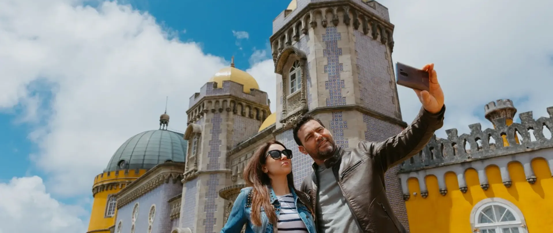 Tourists taking a selfie in front of the colorful and iconic Pena Palace in Sintra, Portugal, under a bright blue sky.