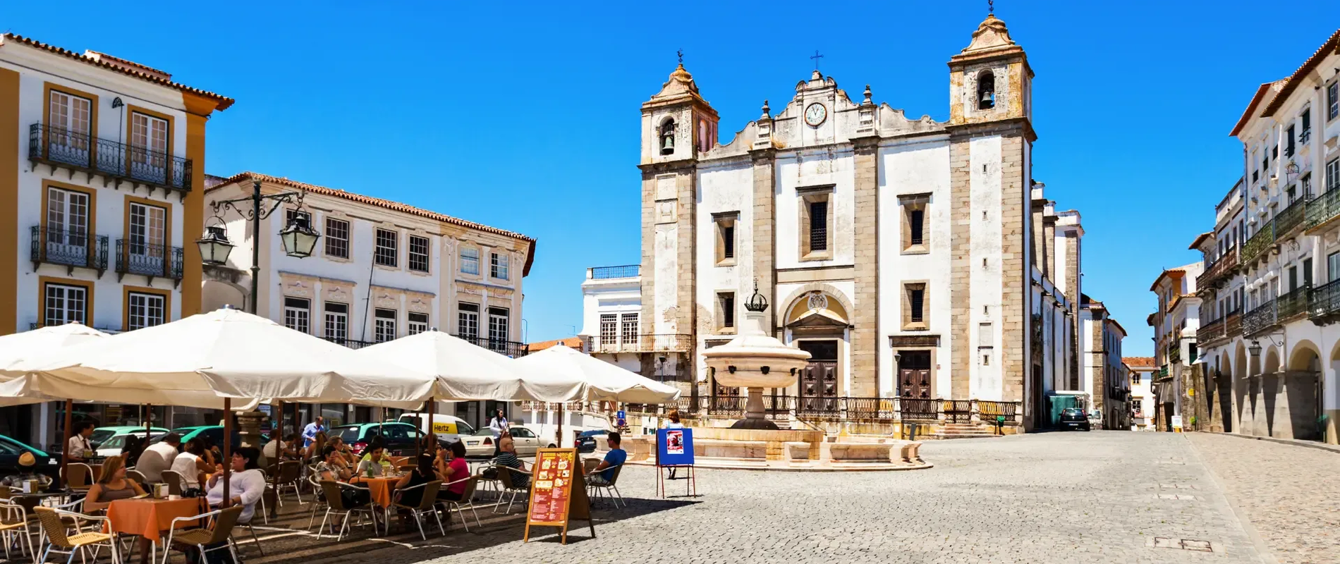 Historic Praça do Giraldo in Évora, Portugal, featuring outdoor cafés, cobblestone pavement, the Santo Antão Church, and the Renaissance fountain under a clear blue sky.
