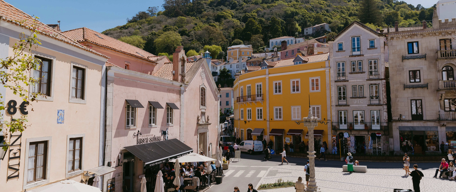 Lively scene from the historic center of Sintra, Portugal, featuring colorful buildings, outdoor cafés, tourists walking, and the Moorish Castle nestled on the green mountain in the background.