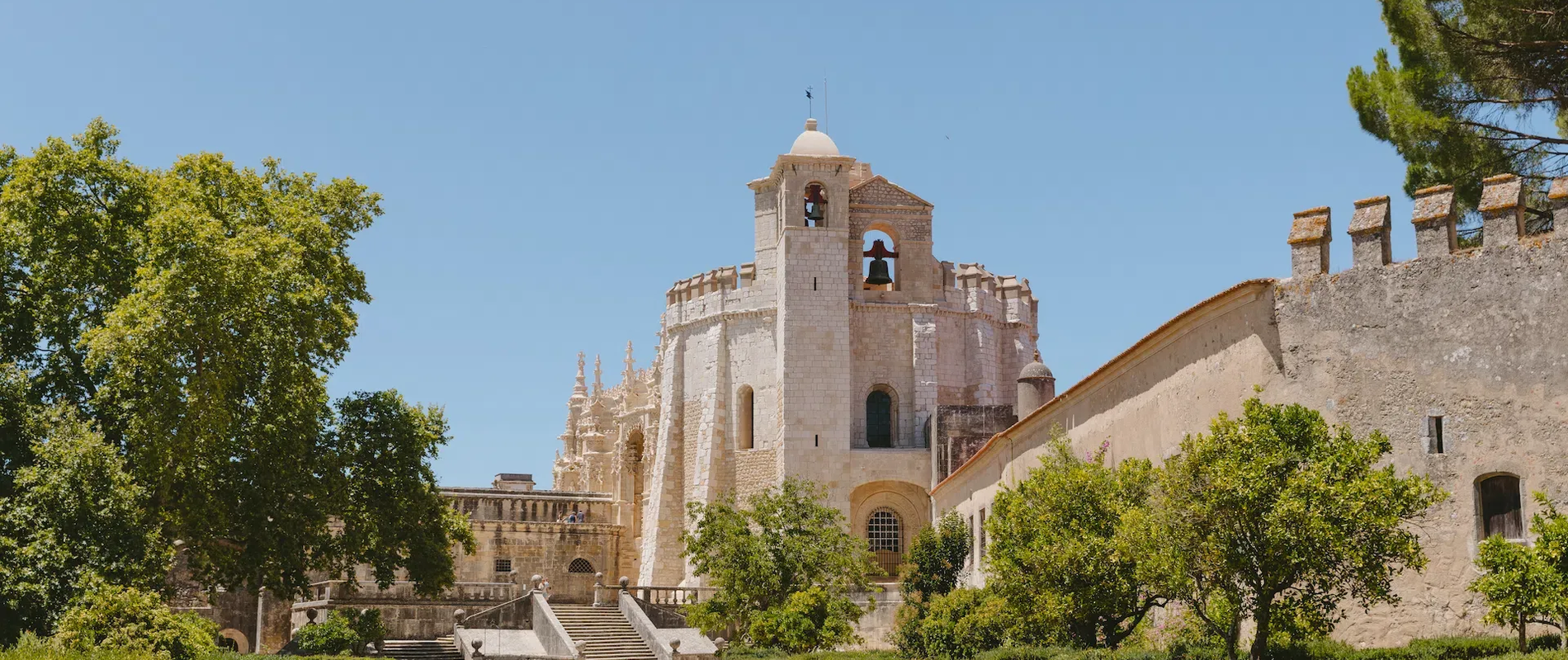 Historic view of the Convent of Christ in Tomar, Portugal, showcasing Manueline architecture and surrounded by lush gardens under a clear blue sky.