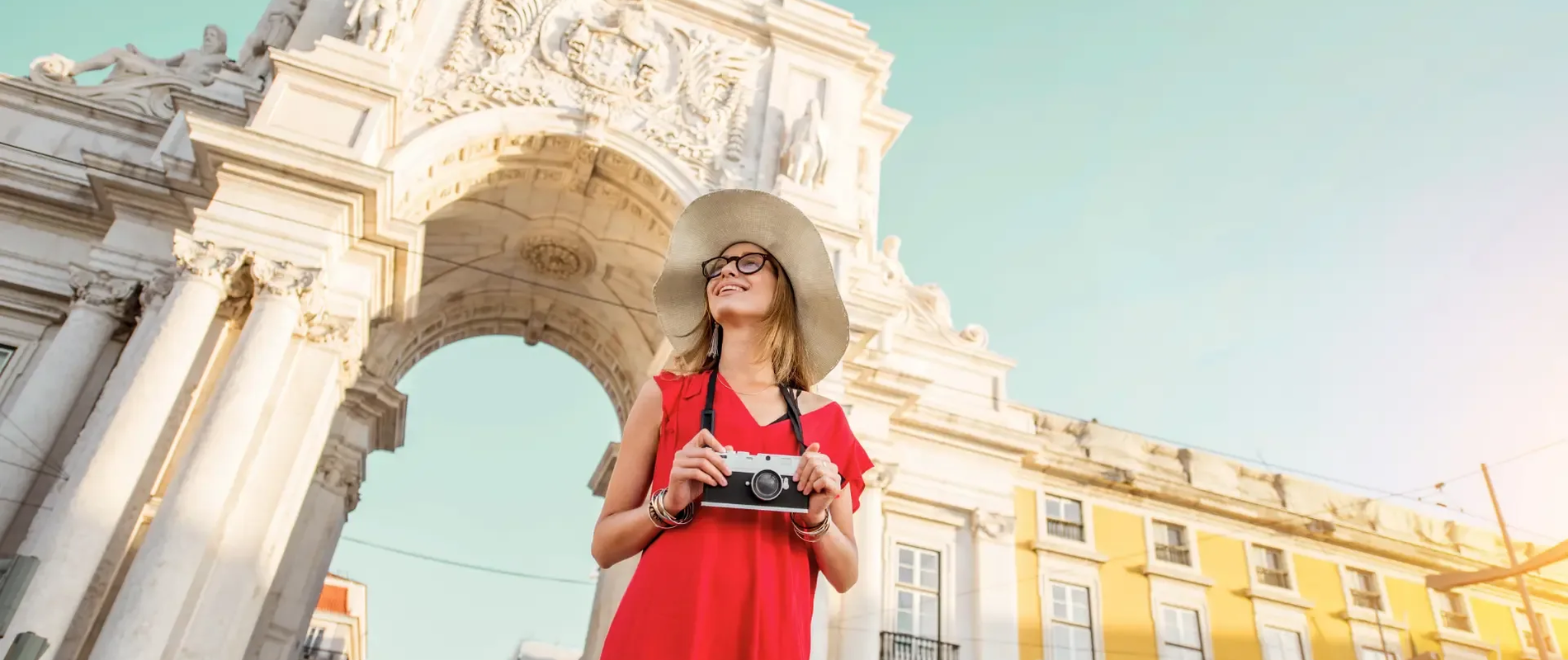Smiling tourist in a red dress and sunhat holding a vintage camera in front of the Arco da Rua Augusta in Lisbon, Portugal.