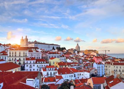Scenic sunset view over Alfama, Lisbon's historic district, featuring red rooftops, the National Pantheon, and São Vicente de Fora Monastery with the Tagus River in the background.