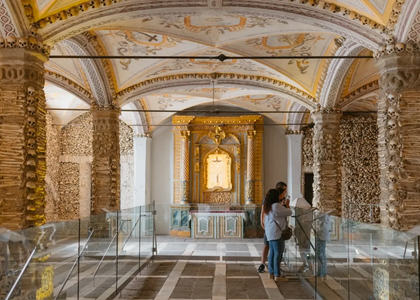 Interior of the Chapel of Bones in Évora, Portugal, with human skulls and bones lining the walls and columns, ornate vaulted ceiling, and visitors observing the golden altar.