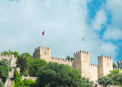 Panoramic view of São Jorge Castle in Lisbon, Portugal, with Portuguese flag and medieval towers under a blue sky.