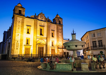 Evening view of Praça do Giraldo in Évora, Portugal, featuring the illuminated Church of Santo Antão and the Renaissance fountain with people relaxing.