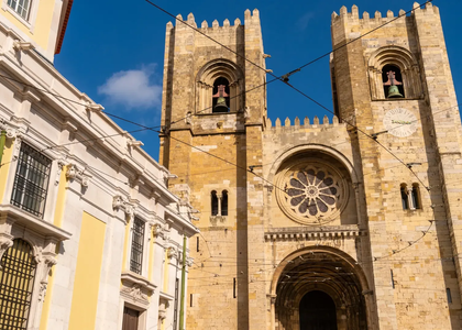 Front view of Lisbon Cathedral (Sé de Lisboa), with its iconic twin towers, rose window, and yellow facade of the adjacent building under a clear blue sky.