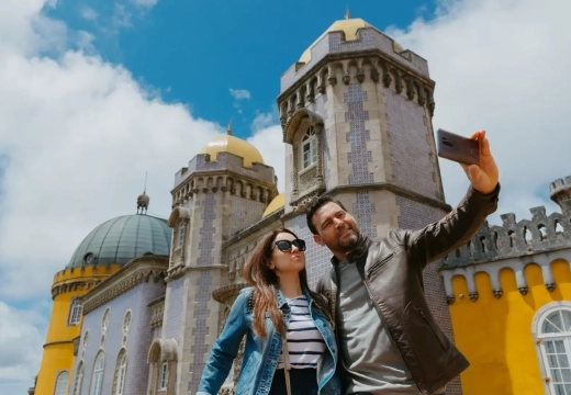 Tourists taking a selfie in front of the colorful and iconic Pena Palace in Sintra, Portugal, under a bright blue sky.