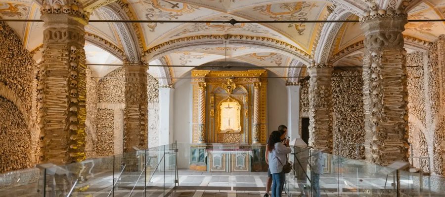Interior of the Chapel of Bones in Évora, Portugal, with human skulls and bones lining the walls and columns, ornate vaulted ceiling, and visitors observing the golden altar.