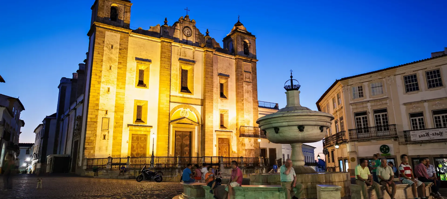 Evening view of Praça do Giraldo in Évora, Portugal, featuring the illuminated Church of Santo Antão and the Renaissance fountain with people relaxing.