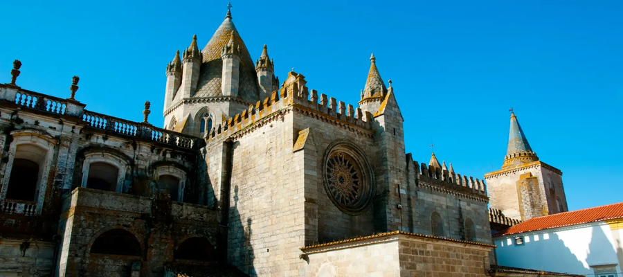 View of the Évora Cathedral in Portugal, a Romanesque-Gothic monument with imposing towers and a rose window under a clear blue sky.