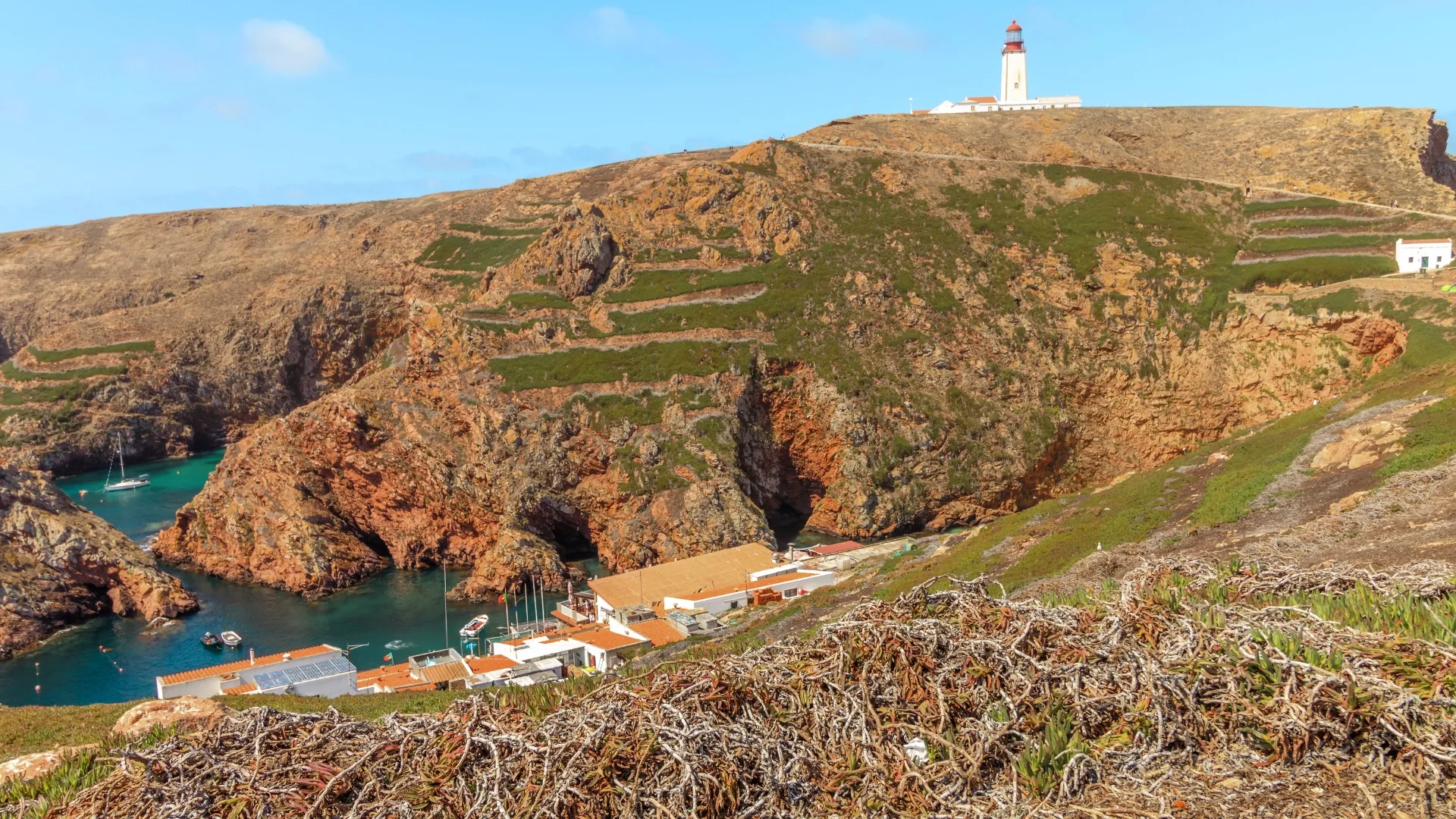 Lighthouse atop Berlenga Island overlooking a dramatic cliffside bay with turquoise waters, boats and traditional white houses nestled by the sea.