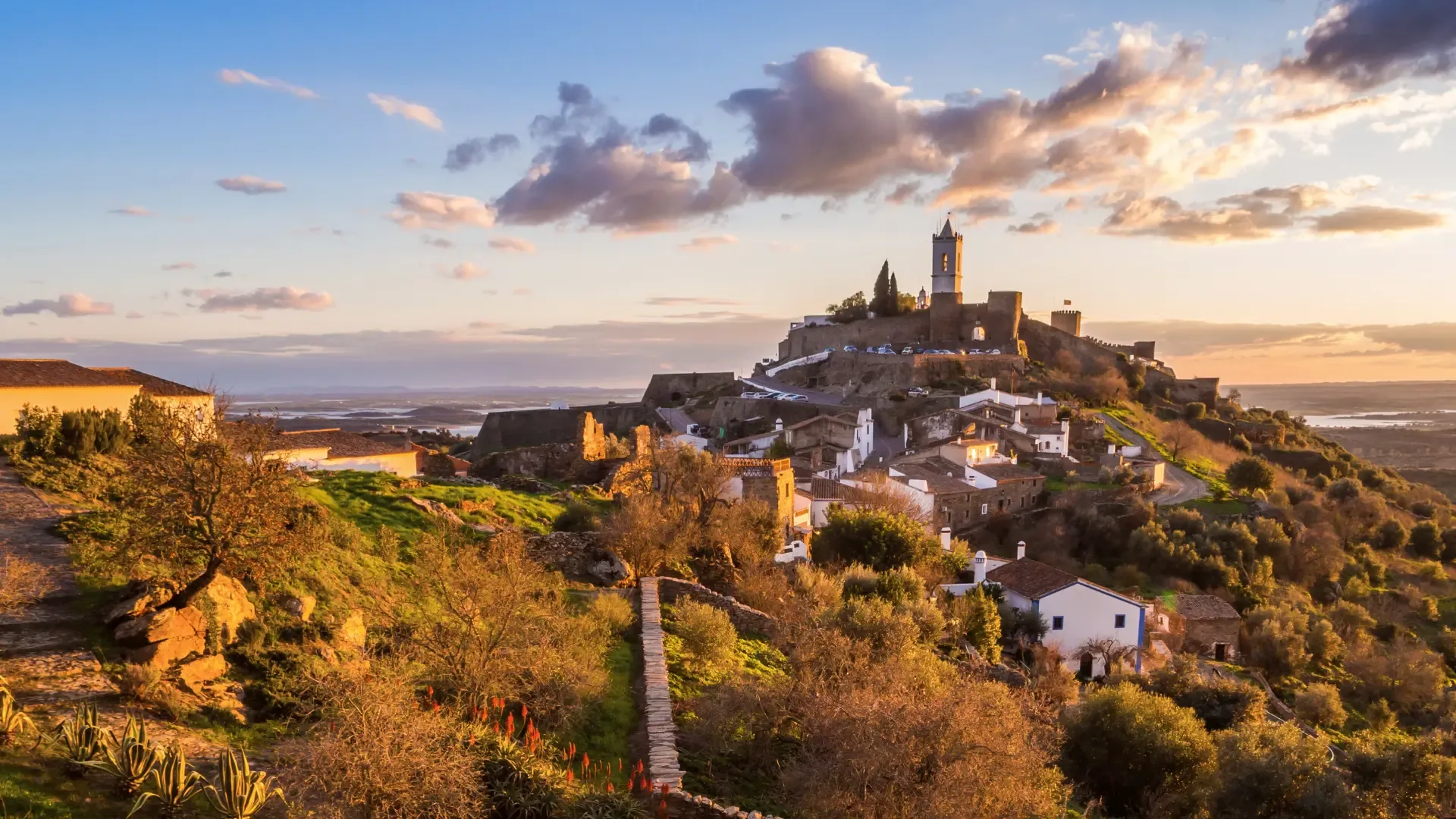 Vista do castelo e vila de Monsaraz ao pôr do sol, com as muralhas, casas caiadas e a paisagem alentejana estendendo-se até ao horizonte.