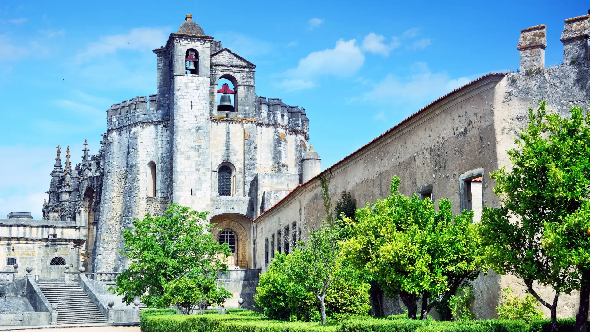 Vista do Convento de Cristo em Tomar, com a sua icónica charola e torre sineira, rodeado por jardins verdes sob um céu azul.