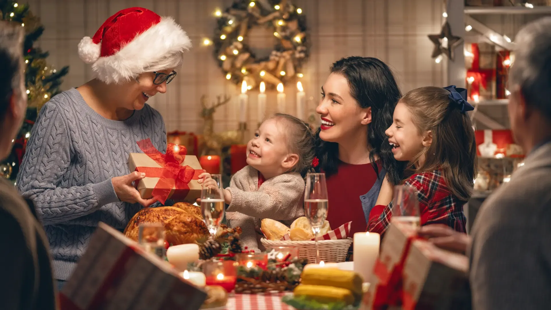 Happy multigenerational family sharing Christmas gifts around a decorated table. A grandmother wearing a Santa hat receives a present from her grandchildren, surrounded by smiles, food, and warm festive lights.