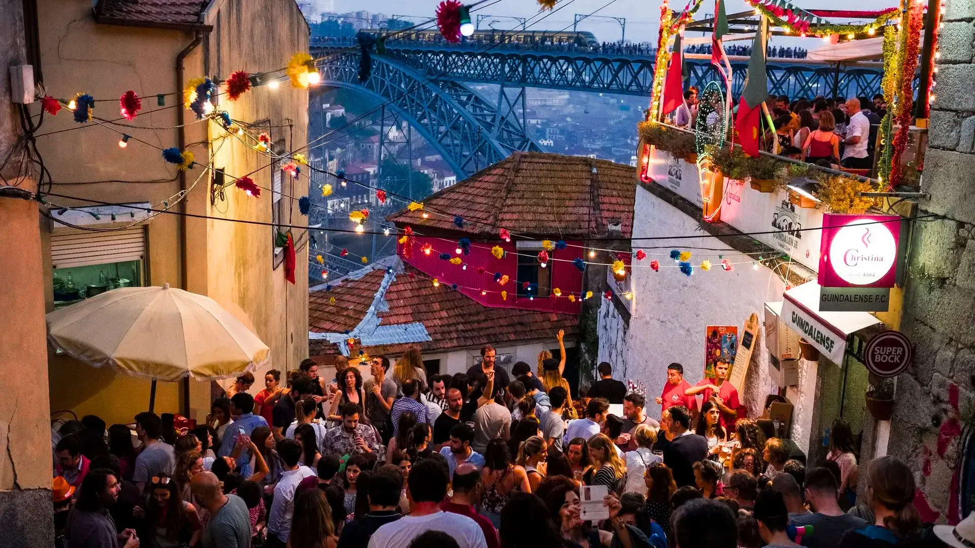 Crowd celebrating São João festival in Porto with colorful decorations, traditional lights and music near the Guindais area, with Dom Luís I bridge in the background.