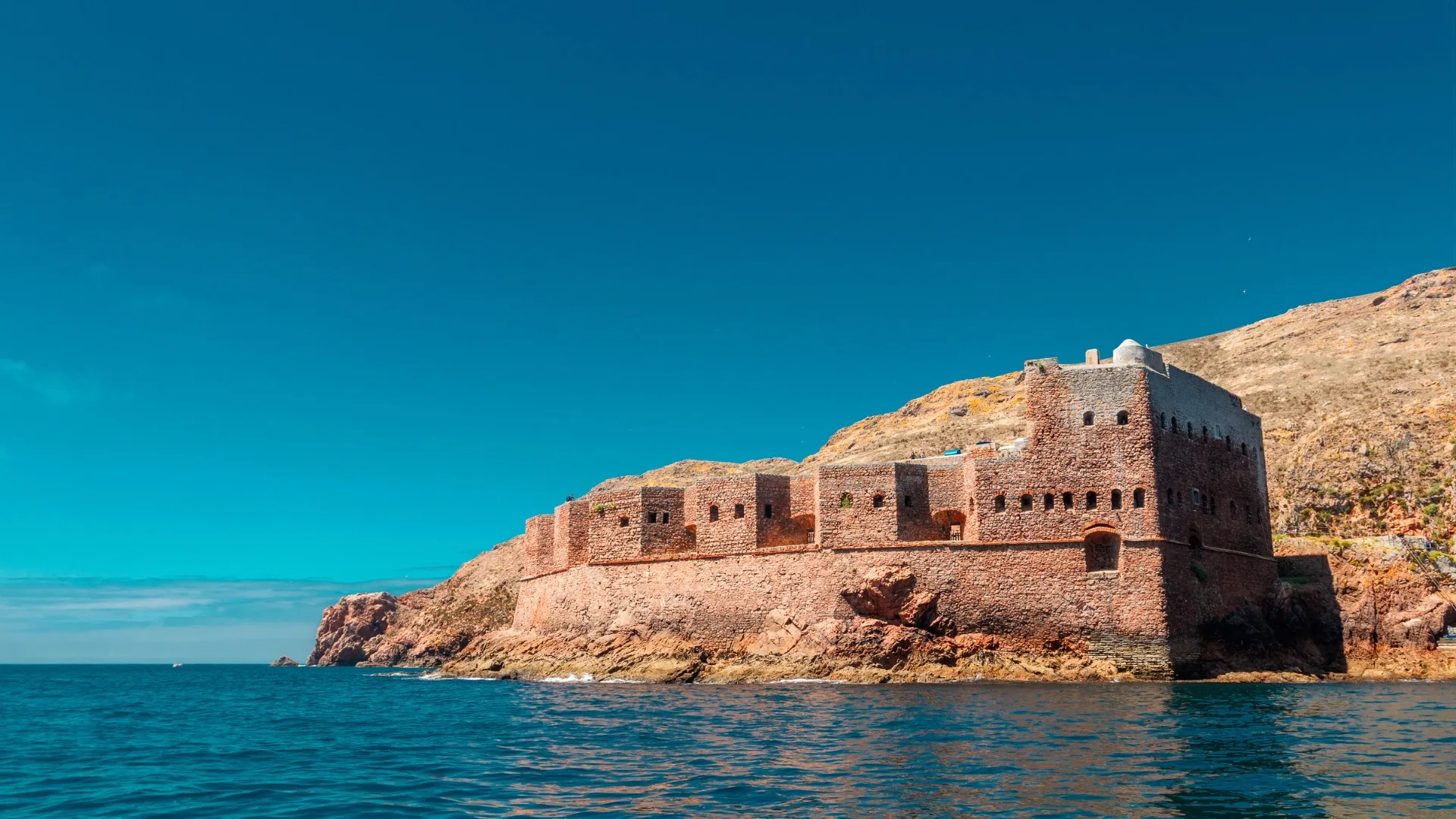Fort of the Berlengas surrounded by turquoise waters and boats, connected to the mainland by a stone path under a bright blue sky.