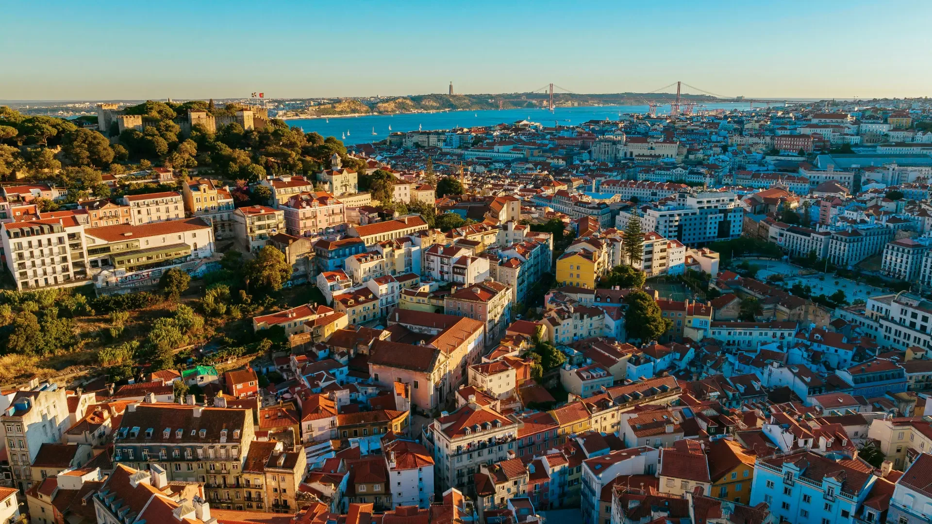 Vista aérea de Lisboa ao final da tarde, destacando o Castelo de São Jorge, o bairro da Alfama e a Ponte 25 de Abril ao fundo, com o rio Tejo iluminado pelo sol.