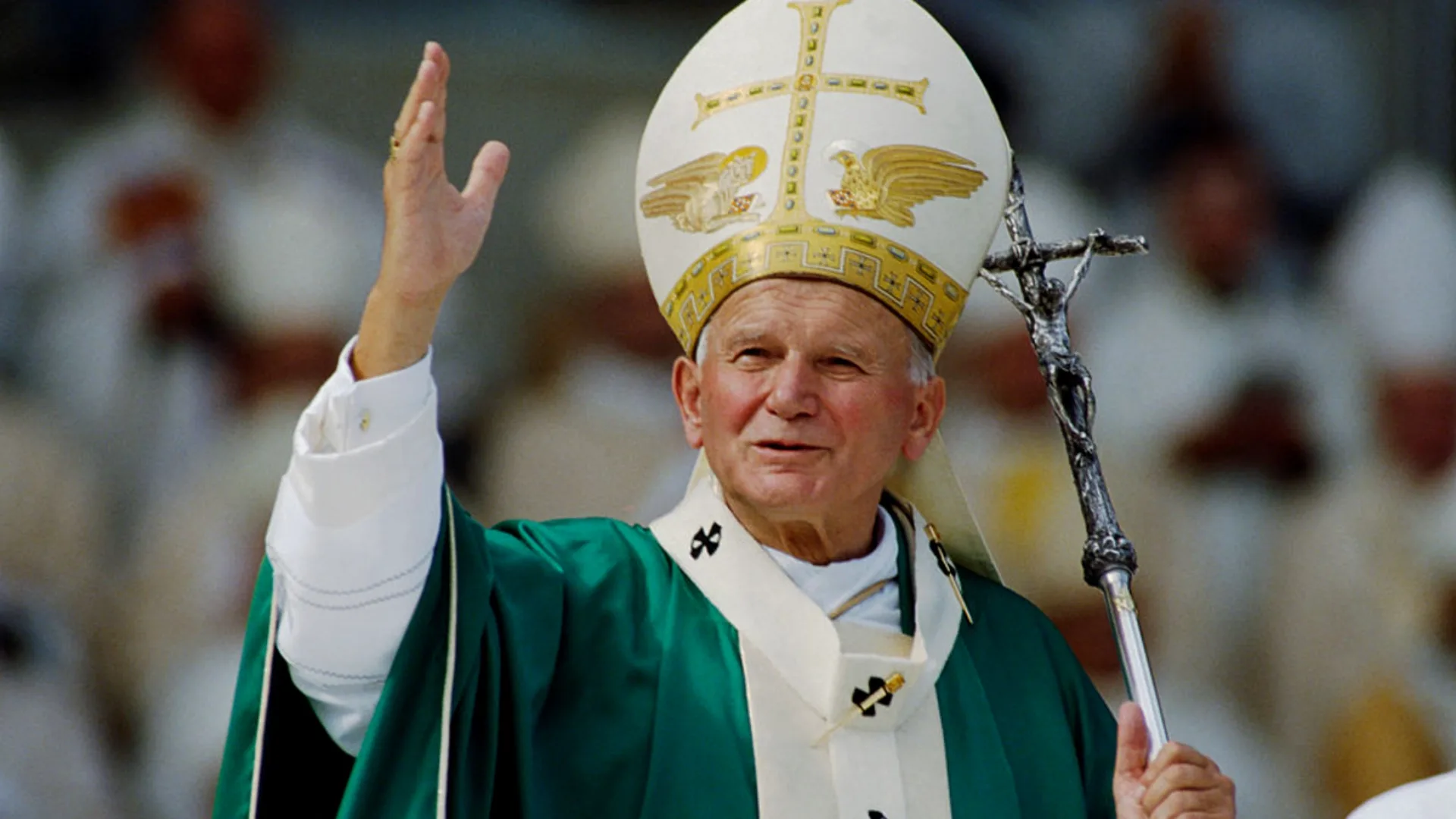 Pope John Paul II blessing the faithful during a mass, wearing green vestments and a mitre, holding a silver papal cross.