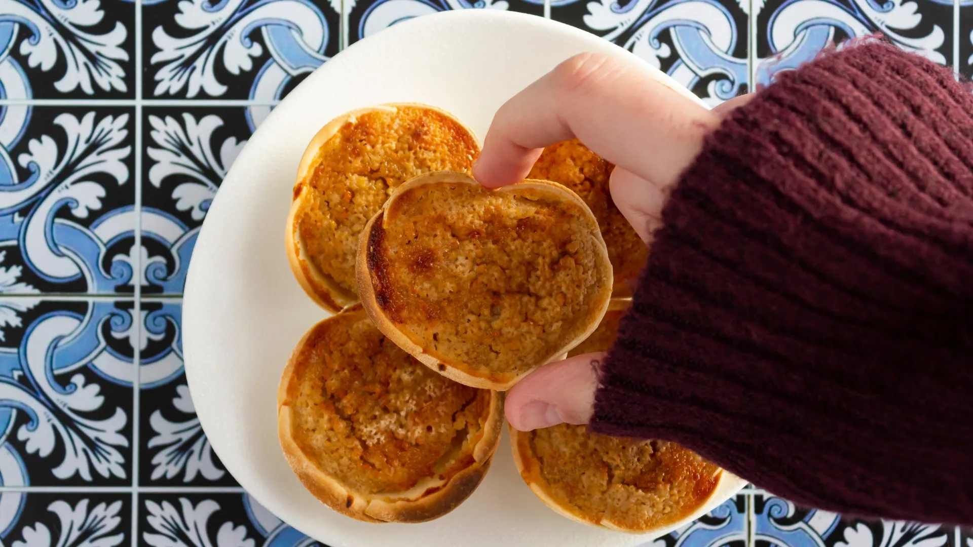 Close-up of traditional Portuguese Queijadas de Sintra on a plate, with a hand picking one, over decorative blue and white tiles.