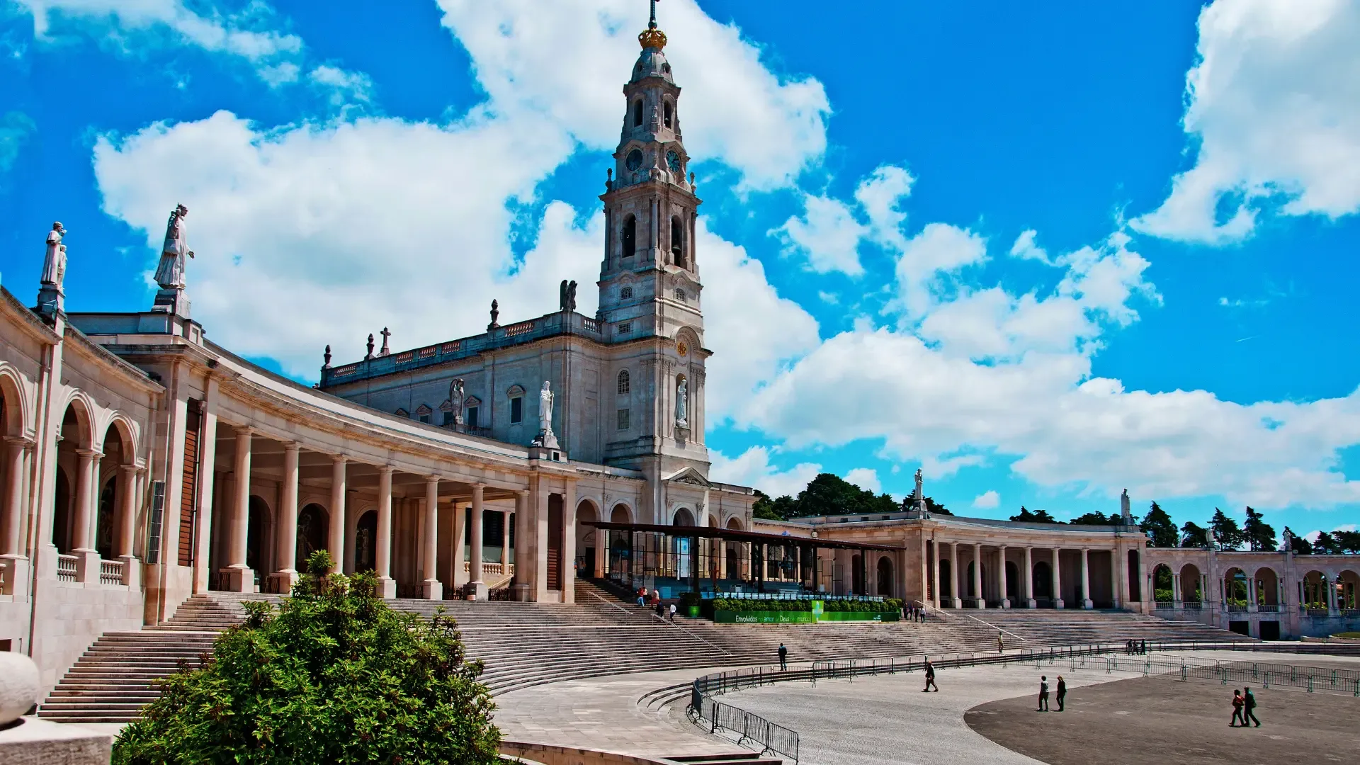 Vista panorâmica do Santuário de Fátima, com a Basílica de Nossa Senhora do Rosário sob um céu azul com nuvens e a esplanada em primeiro plano.