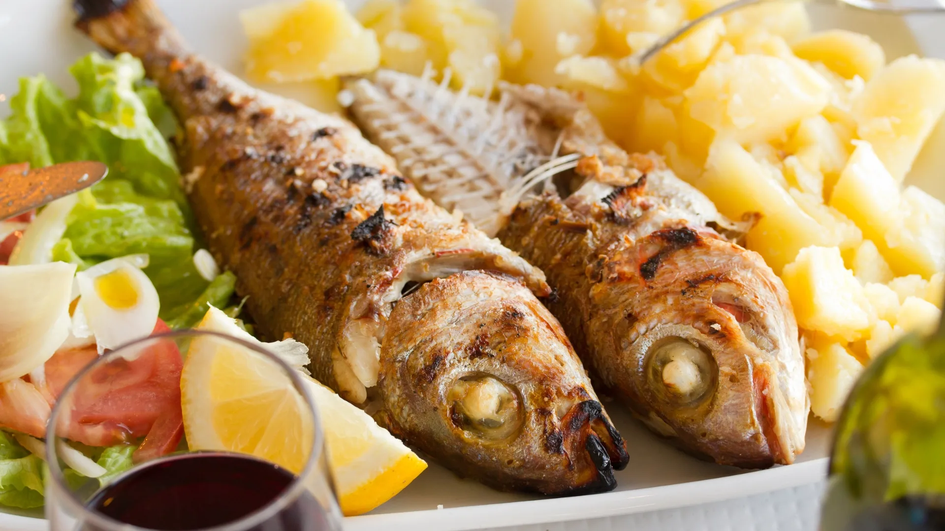Close-up of traditional Portuguese grilled sardines served with boiled potatoes, fresh salad, lemon, and a glass of red wine.