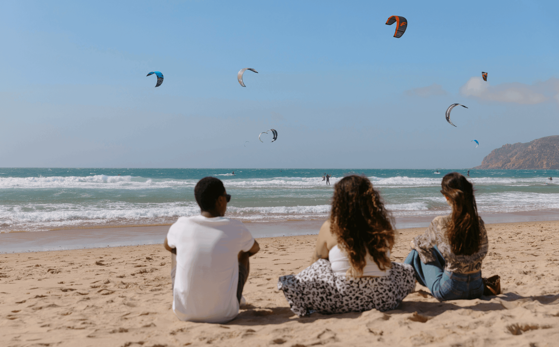 Three friends sitting on the sand watching colorful kitesurfers glide over the ocean waves on a sunny beach in Portugal. The scene captures a relaxed, adventurous moment by the Atlantic coast.