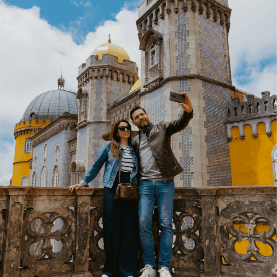Couple taking a selfie at the colorful Pena Palace in Sintra, Portugal. The UNESCO World Heritage site features vibrant yellow and blue towers, with romantic and neo-Gothic architecture set against a partly cloudy sky.