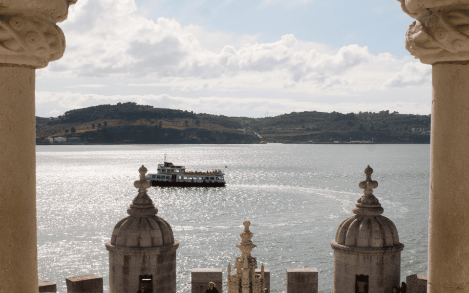 Framed view from the Belém Tower in Lisbon, Portugal, showing a traditional ferry crossing the shimmering waters of the Tagus River. Manueline-style architectural details in the foreground highlight the historic monument.