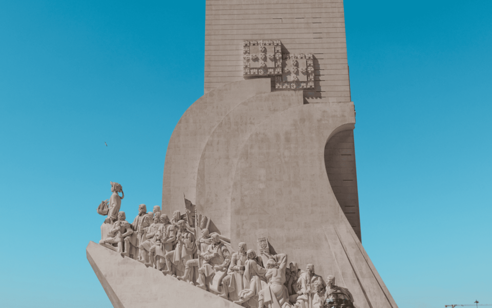 Side view of the Monument to the Discoveries (Padrão dos Descobrimentos) in Belém, Lisbon, Portugal. The statues of Portuguese explorers, monarchs, and visionaries are prominently displayed against a bright blue sky.