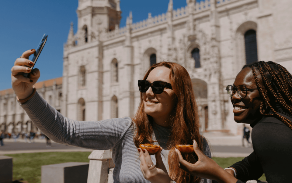 Two smiling women taking a selfie while enjoying Pastéis de Belém in front of the Jerónimos Monastery in Lisbon, Portugal. A joyful travel moment combining local pastry, heritage, and culture.