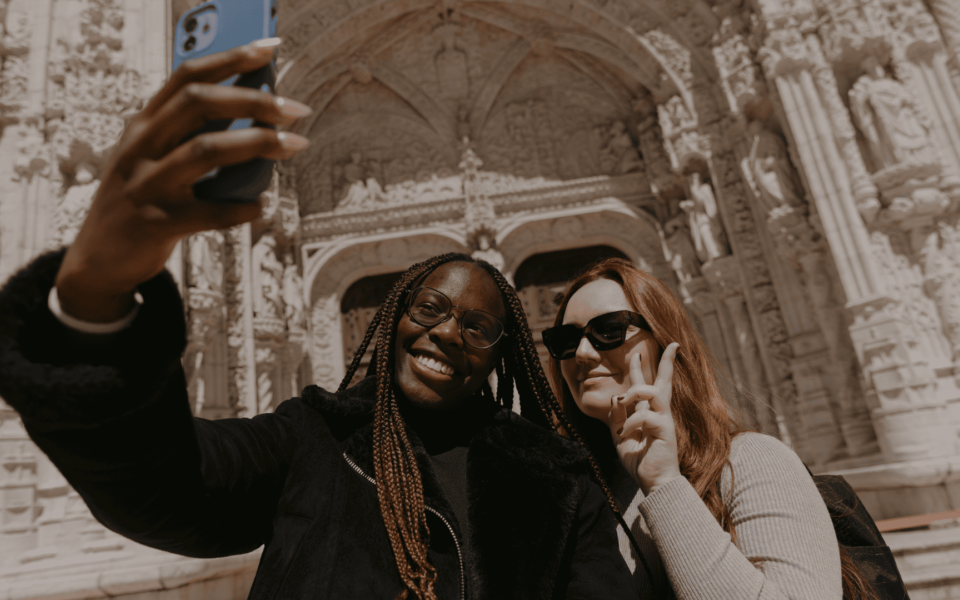 Two smiling women taking a selfie in front of the ornate Manueline entrance of the Jerónimos Monastery in Lisbon, Portugal. A cultural heritage experience combining history and modern tourism.