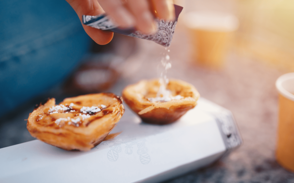 Close-up of a person sprinkling powdered sugar over two freshly baked Pastéis de Belém, the iconic Portuguese custard tarts, placed on a branded box with warm sunlight in the background.