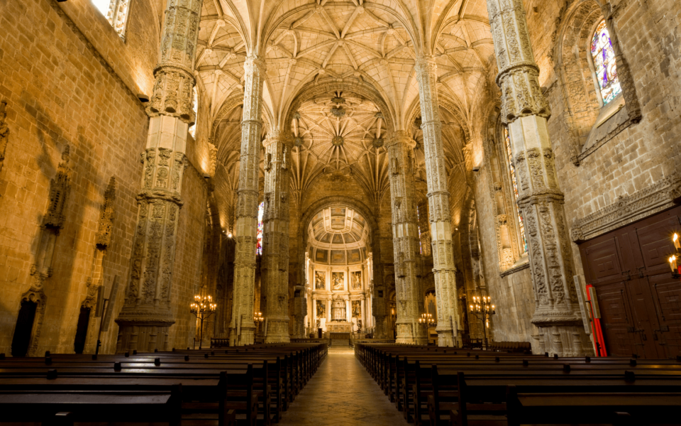 Majestic interior view of the Church of the Jerónimos Monastery in Lisbon, Portugal, showcasing intricate Manueline architecture, ornate stone columns, ribbed vaults, and the grand altar at the far end.