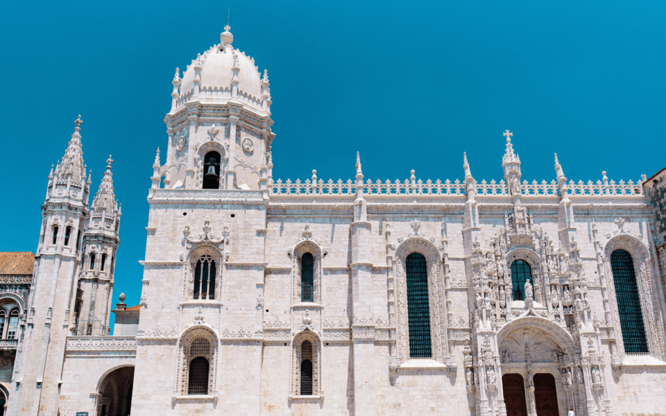 Front view of the south façade of the Jerónimos Monastery in Lisbon, Portugal, featuring elaborate Manueline architecture, sculpted arches, and towering spires against a clear blue sky.