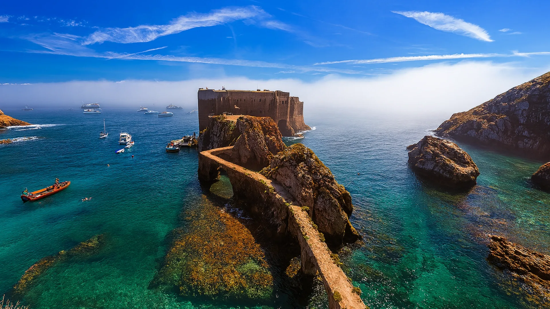 Historic São João Baptista Fort connected by a stone bridge over turquoise waters on Berlenga Island, surrounded by cliffs and anchored boats.