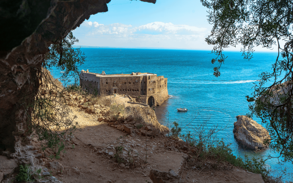 Framed view of the Fort of São João Baptista on Berlengas Island, Portugal, seen through natural cliffs and vegetation. The fortress overlooks the deep blue Atlantic Ocean, with a boat approaching from the sea.