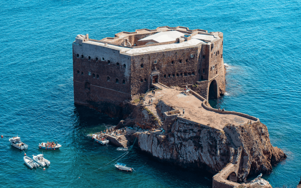 Aerial close-up of the Fort of São João Baptista on Berlengas Island, Portugal, highlighting the rocky access path, main gate, and boat landing platform surrounded by clear Atlantic waters and small visitor boats.