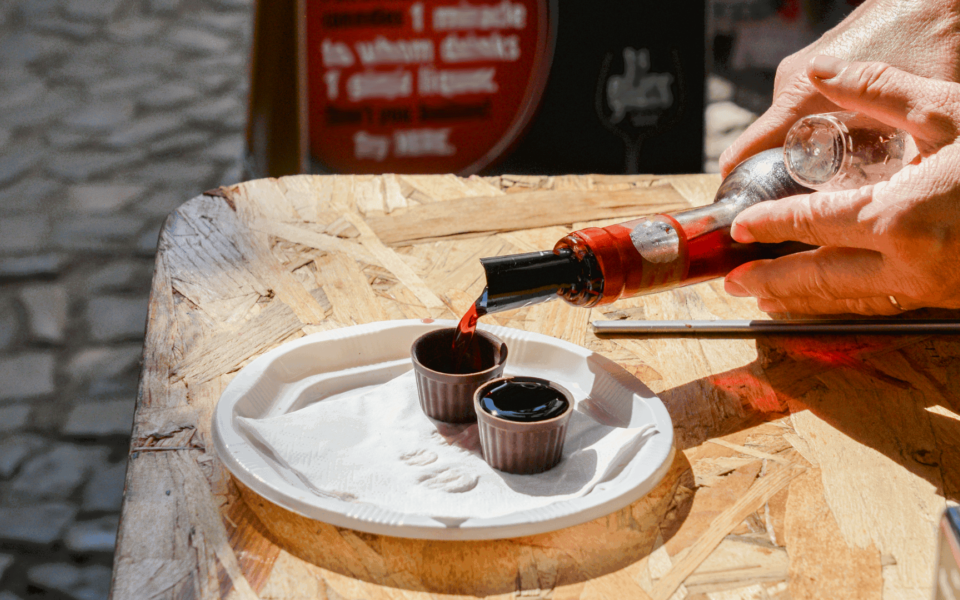 Close-up of a person pouring Ginjinha, the traditional Portuguese cherry liqueur, into small chocolate cups during a tasting session in Óbidos. The setup is on a rustic wooden counter under natural sunlight.