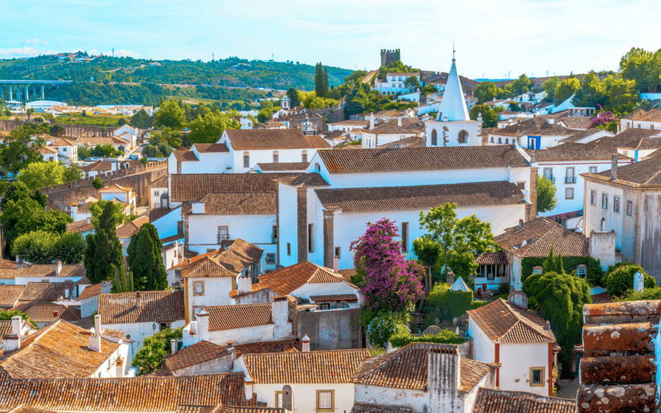 View over the traditional whitewashed houses and terracotta rooftops of Óbidos, Portugal, with vibrant greenery, blooming flowers, and the castle walls in the distance. A picturesque historic village surrounded by natural beauty.