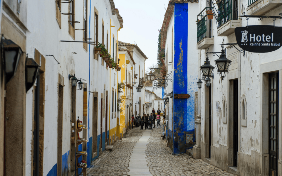 Typical cobblestone street in the medieval village of Óbidos, Portugal, lined with whitewashed houses, colorful façades, hanging lanterns, and a local hotel sign. Visitors walk through the charming historic alley.