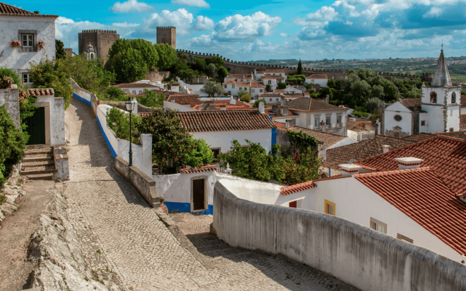 Scenic view of the medieval village of Óbidos in Portugal, showcasing cobblestone streets, whitewashed houses with red-tiled roofs, and the historic castle walls surrounding the town. A popular cultural and historical destination.