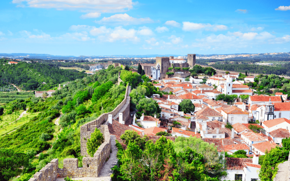 Panoramic aerial view of the medieval walled town of Óbidos in Portugal, featuring red-roofed houses, historic churches, and the well-preserved castle surrounded by lush green hills and fortified stone walls.