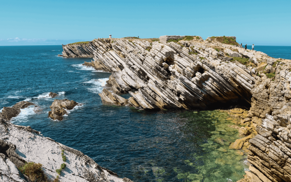 Striking coastal cliffs in Peniche, Portugal, with layered rock formations sloping toward the Atlantic Ocean. The clear blue and green waters highlight the natural beauty of Portugal's west coast and its unique geological landscape.