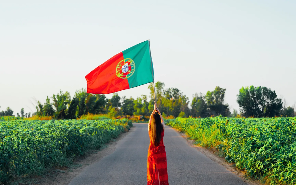 Woman waving the Portuguese flag on a rural road surrounded by green fields and trees under a clear sky.