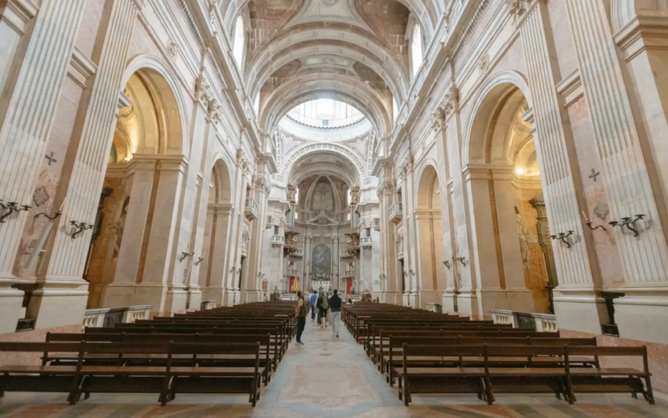 Interior view of the Basilica of the Mafra National Palace, showcasing the grand marble nave, high vaulted ceilings, and baroque architectural details.