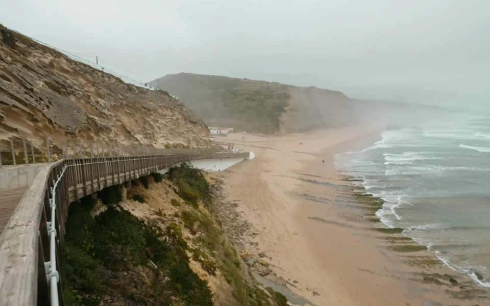 Wooden walkway along the cliffs of Santa Rita Beach in Torres Vedras, Portugal, with misty waves crashing on the sandy shore.